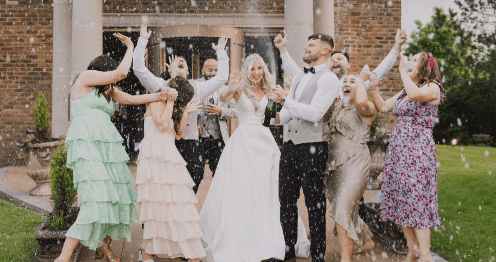 A joyful wedding celebration with a bride, groom, and guests popping champagne outside a brick manor venue, all dressed in formal attire, smiling and laughing.